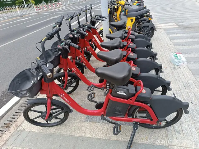 The image shows a row of red and black e-bikes parked on the side of a street, with a white pole in...