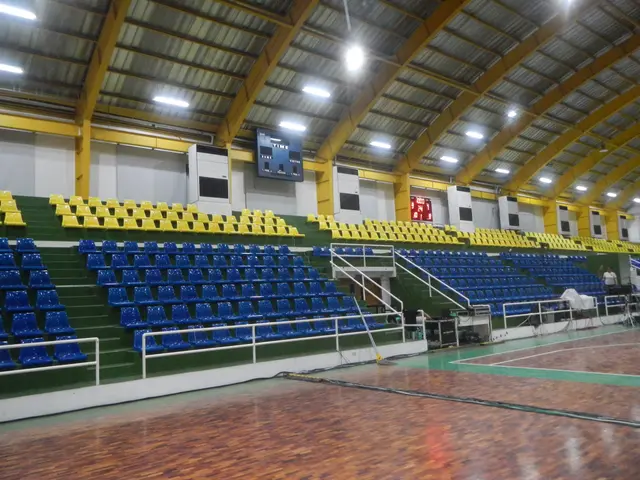 The image shows a large indoor sports hall with a wooden floor, blue and yellow chairs, railings,...