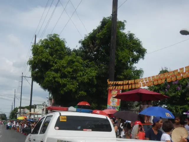 The image shows a police truck driving down a street with a group of people standing on the side of...