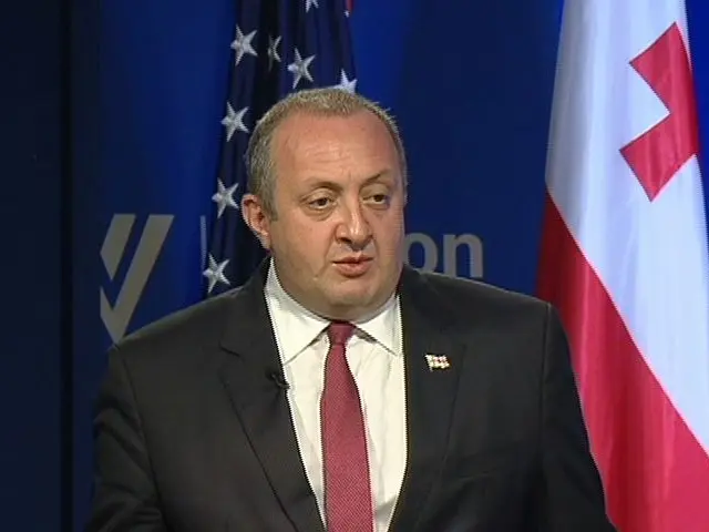 The image shows a man in a suit and tie standing at a podium with two flags behind him and a blue...