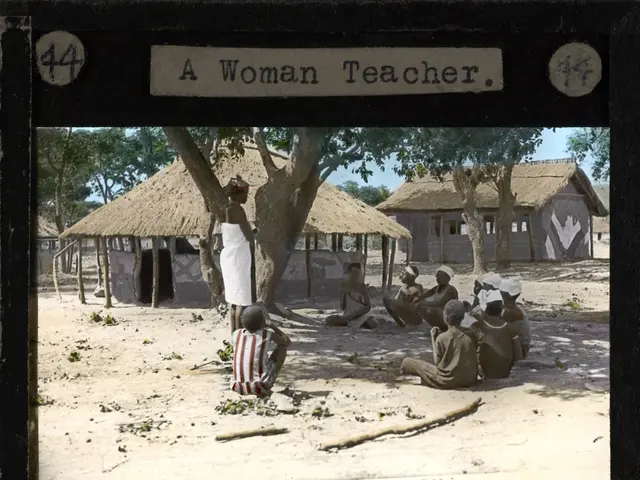 The image shows a woman teacher standing in front of a group of people sitting on the ground, with...
