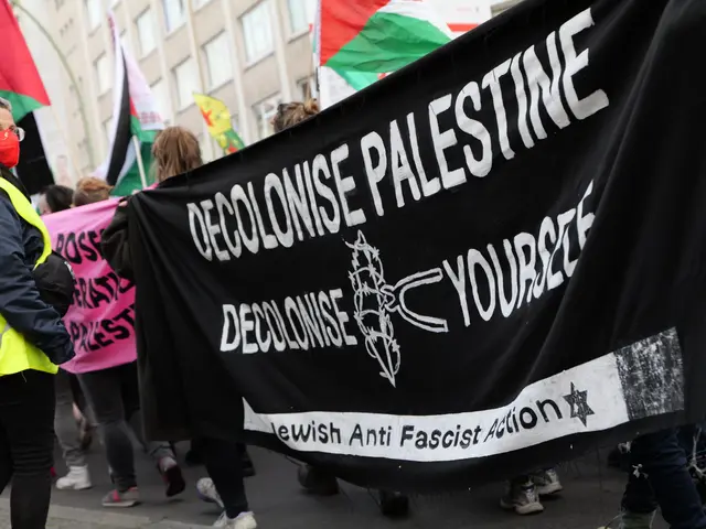 The image shows a group of people walking down a street, holding a black banner with the words...