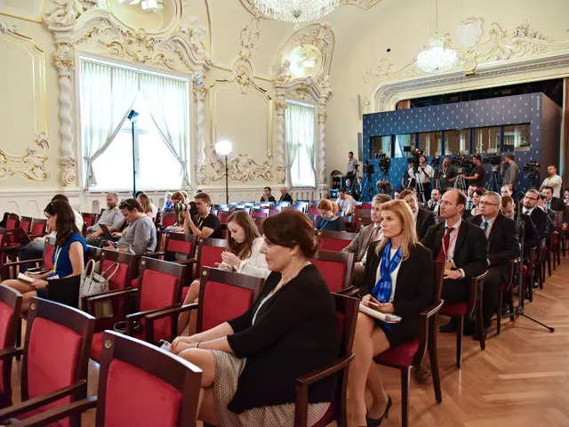 The image shows a large group of people sitting in chairs in a room with a wall in the background,...