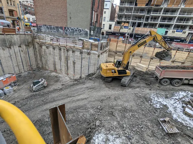 The image shows a construction site with a large excavator in the middle of it, surrounded by...