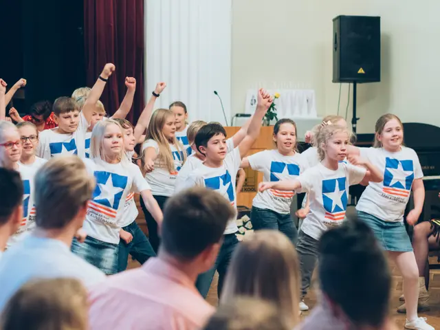The image shows a group of children in white shirts and blue shorts dancing in front of a crowd of...