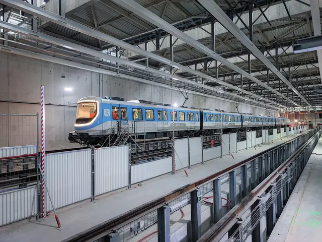 The image shows a blue and white train traveling through a large building, with a platform on the...