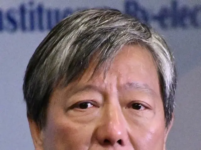 The image shows a man in a suit and tie speaking into a microphone in the foreground, while in the...